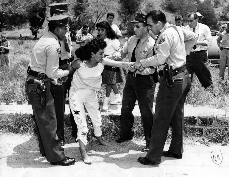 Archival black-and-white photo of four sheriff's deputies gripping the wrists of a barefoot woman as she resists.